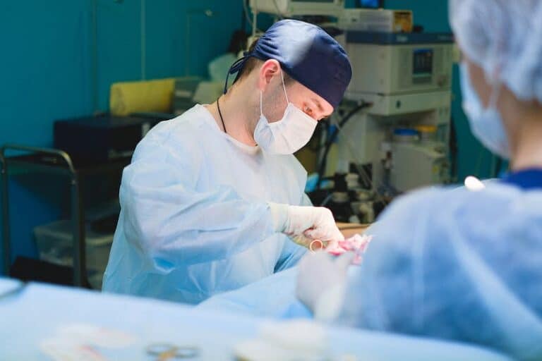 Surgeon in a blue cap and mask operating with an assistant in the background